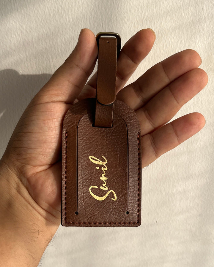 A hand holding a brown textured leather luggage tag with a gold script logo that reads 'Snevil' against a white wall with soft shadows.